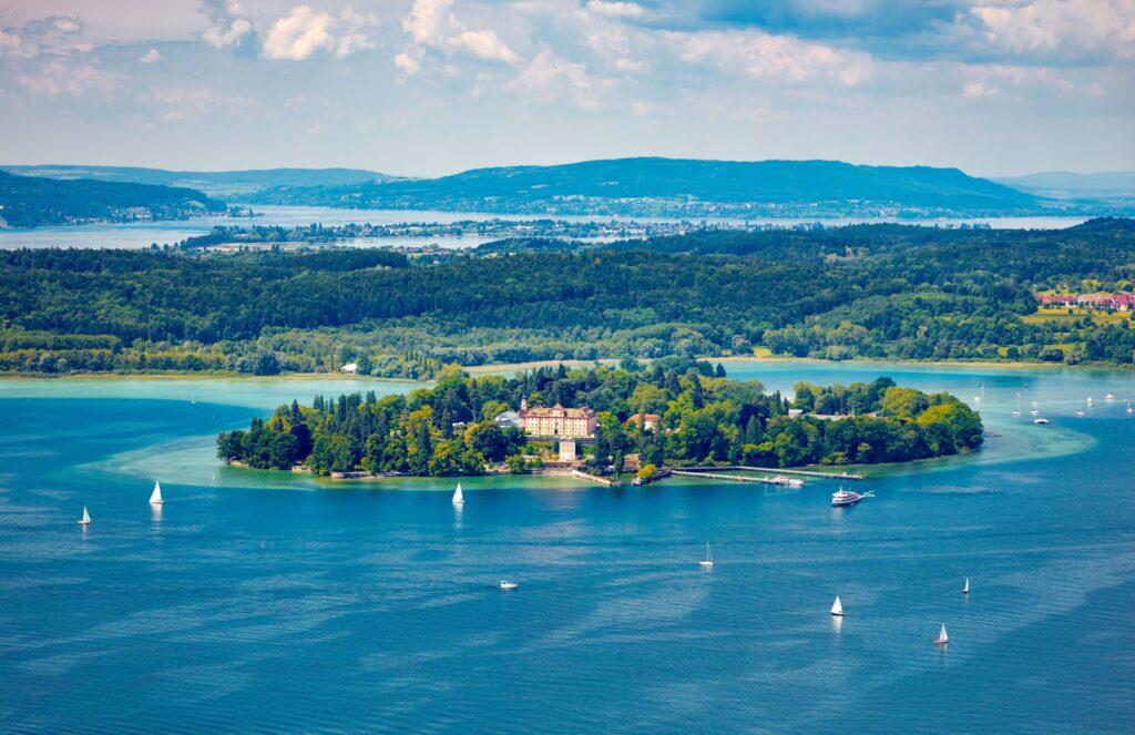 Insel Mainau im Bodensee – Luftaufnahme der Insel Mainau mit ihrem Schloss, umgeben von blauem Wasser und Segelbooten, mit einer weiten Aussicht über die Bodensee-Region.